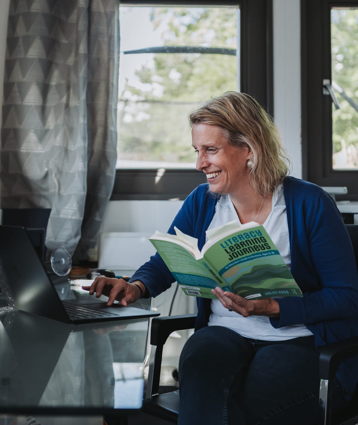 Dr Helen Ross laughing at her laptop, holding her book Literacy Learning Journeys